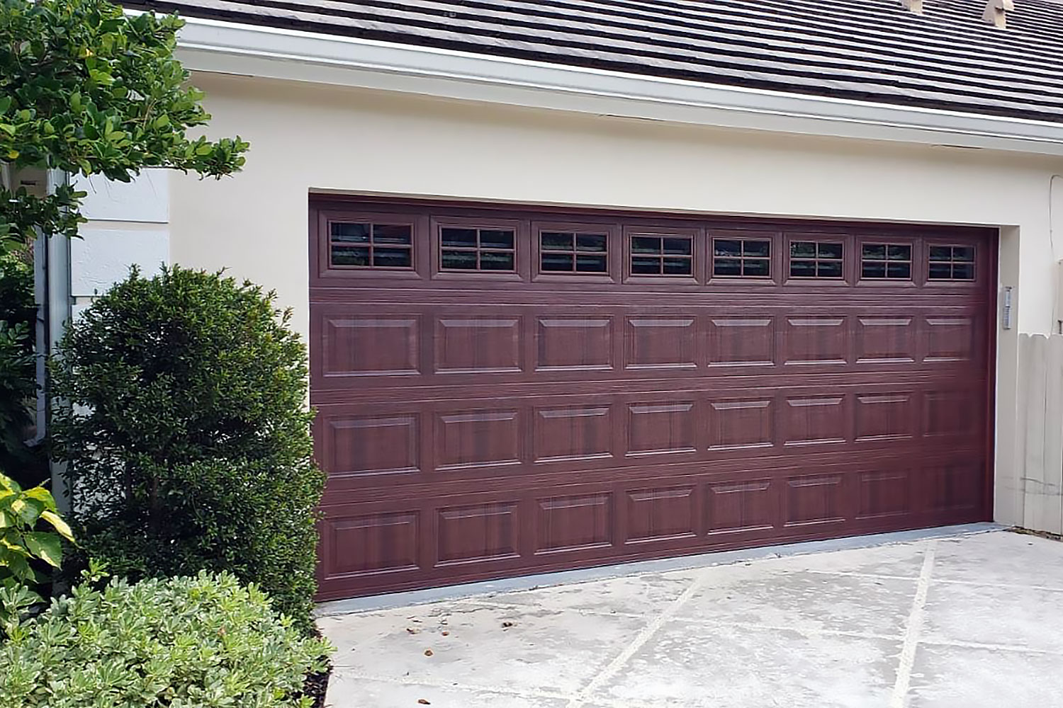 A burgundy sectional garage door with rectangular panels, flanked by shrubs and a concrete driveway.